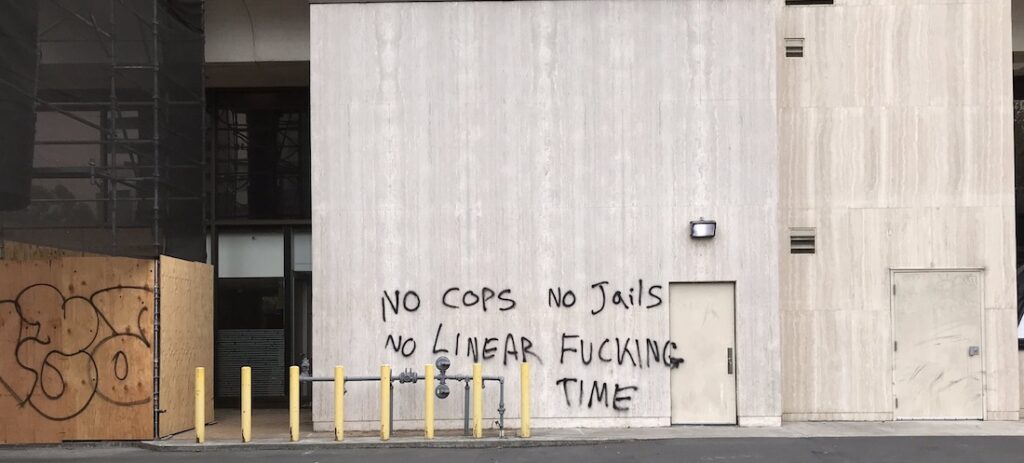 Graffiti on wall next to boarded building, reading: "No cops no jails, no linear fucking time."
