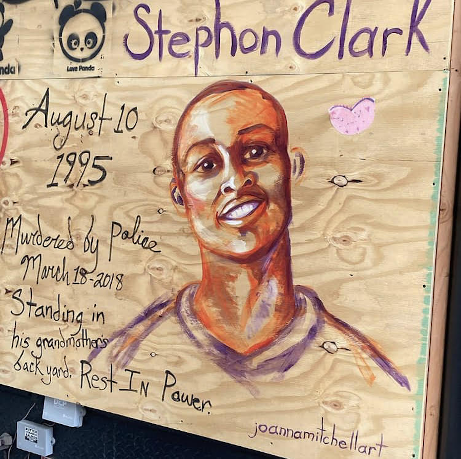 A wooden board over a storefront window is painted over with a detailed portrait of a young Black man’s smiling face and a pink heart beneath the name Stephon Clark. To the left of the portrait are the words “August 10 1995 Murdered by police March 18 2018 Standing in his grandmother’s backyard. Rest In Power.” Portrait artist JoAnna Mitchell in Soho, New York City.