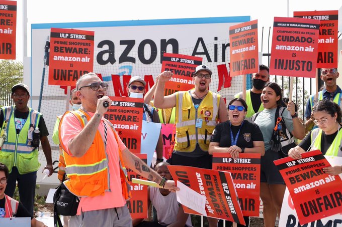 A man in an orange vest, surrounded by picketers holding red picket signs with their fists raised, speaks to the crowd with a microphone.