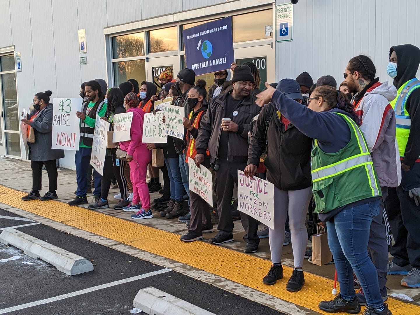 A crowd of workers wearing reflective vests and supporters in coats and hats stand in front of the glass entrance to Amazon Delivery Station DMD9, facing the parking lot and holding hand-made signs that read, “$3 RAISE NOW!” “AMAZON ESCUCHA ESTAMOS EN LA LUCHA!” “AMAZONIANS UNITED,” “WHERE’S OUR $3,” “When we fight back we win,” “AMAZON IS NOTHING WITHOUT ITS WORKERS,” and “JUSTICE FOR AMAZON WORKERS.”