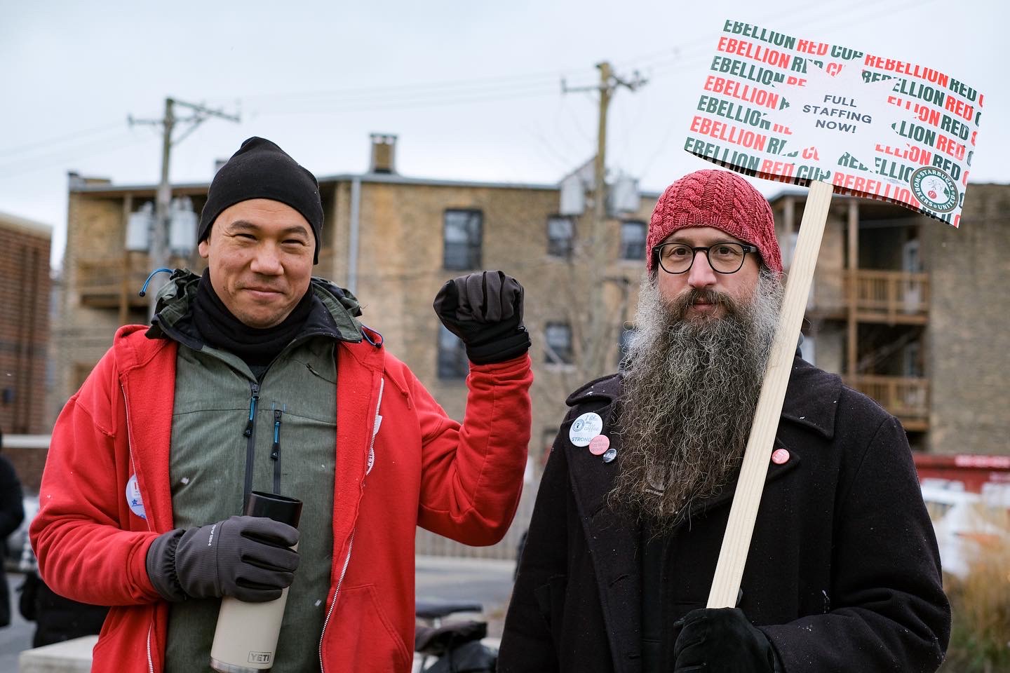 A supporter holds up a begloved fist in solidarity next to a picketer with a long salt and pepper beard holding a “FULL STAFFING NOW” sign.