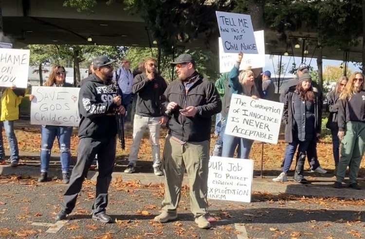 A crowd of protesters, most wearing jeans, stands along a sidewalk. One protester holds a long pole shaped like a diamond-shaped orange caution sign that reads “Save Our Children.” Two other protesters hold signs that read “SHAME ON YOU” and “SAVE GOD’S CHILDREN!”