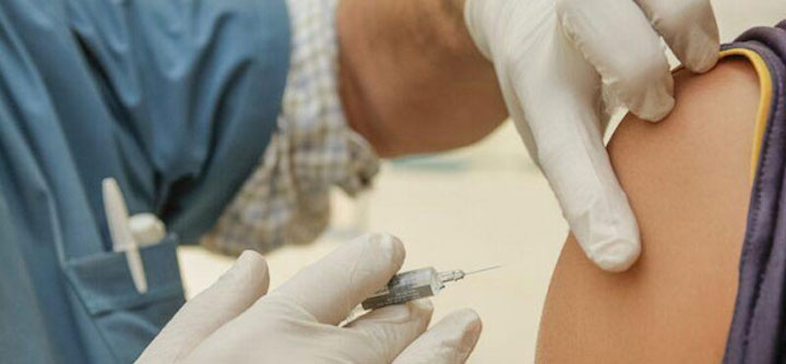 A medical worker in blue prepares to inject a person's shoulder.