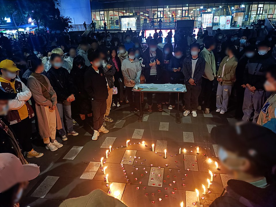 A large group of students of Southwest Jiotong University stand outside a university hall, in front of a table, and surrounding a makeshift altar, made out of burning candles in the shape of a heart, to mourn the victims of the Urumqi fire.