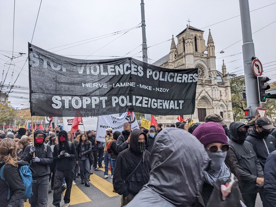 A good size crowd of protestors is marching on a street in Geneva, Switzerland in front of a large church and near a traffic light. Some of them are holding signs including a few people carrying a large black banner with white text that reads across three lines the following: Stop Violences Policieres in French on the first line (translated to Stop Police Violence in English) Herve, Lamine, Mike, Nzoy on the second line (the names of four Black men killed by Swiss police) Stoppt Polizeigewalt in German on the third line (translated to Stop Police Violence in English.