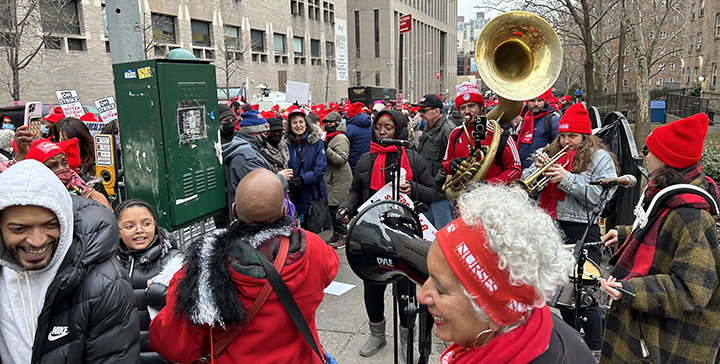 A lively and bustling picket line takes place on the sidewalks and street of Madison Ave in front of Mount Sinai hospital. In the forefront, a band plays music with one person singing into a microphone, another playing tuba, another playing trumpet, and someone drumming a single snare. Several people are smiling or laughing as they stand in front of the band, one of whom is wearing a red headband that says “NURSES” and is holding a megaphone. Many people stand in the background, wearing NYSNA red hats and holding various signs that say “ON STRIKE.”