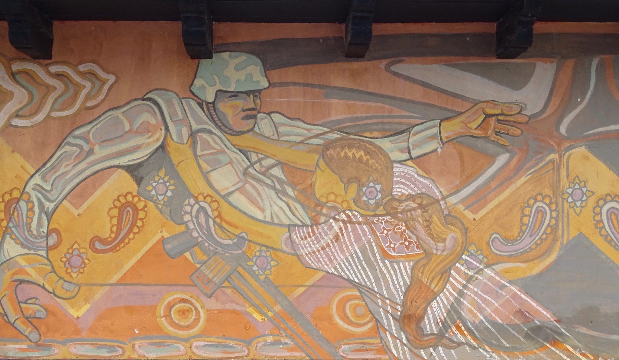 A mural painted onto a wall shows a woman attacking a taller and bigger soldier. The woman is wearing Indigenous Mexican clothing with a long ponytail and a flower in her hair. The soldier is wearing a military uniform and helmet, and has a rifle strapped around his soldier. He seems to be defeated by the woman, with his hands floating as he gets pushed down by her. The mural is made up mostly of oranges, yellows, pinks and grays. Several patterns are painted into the mural.