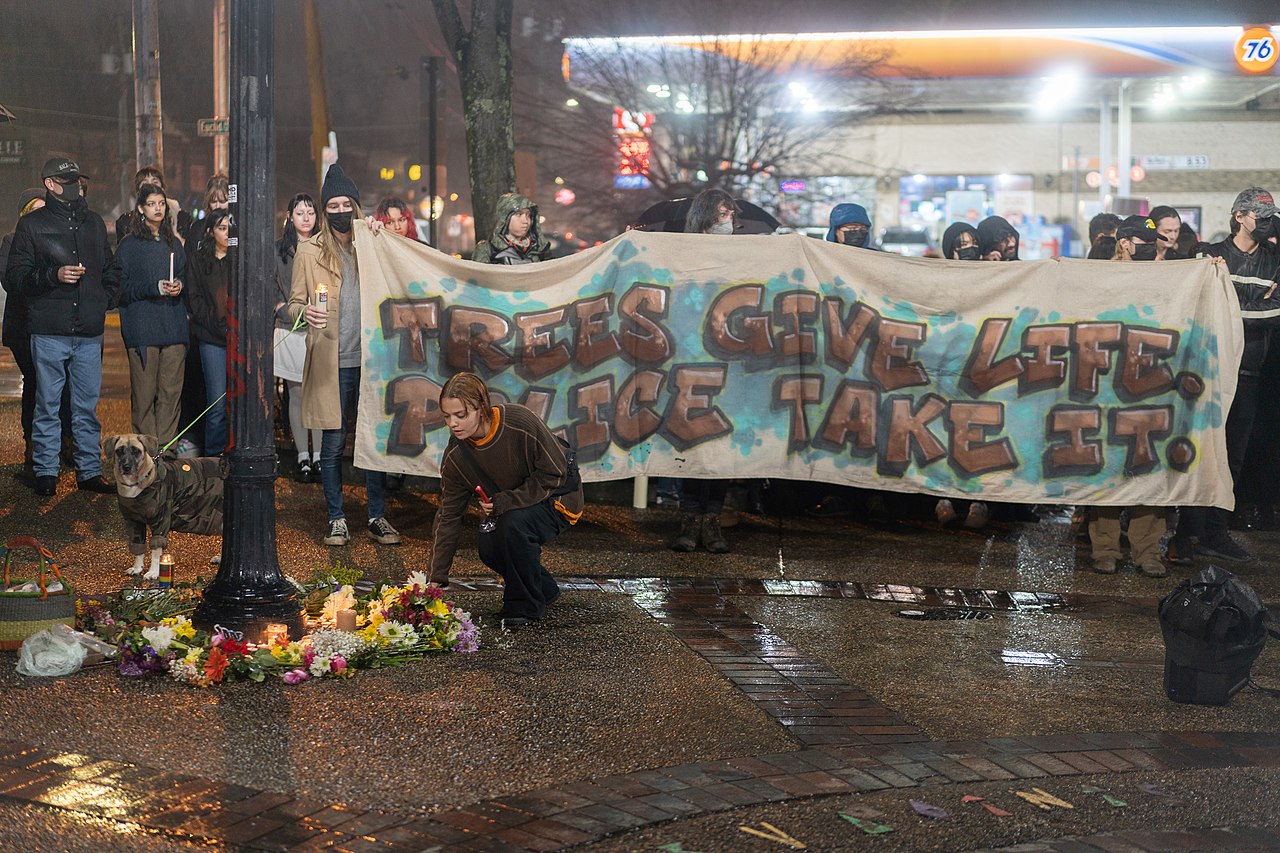 A shrine of bouquets of flowers, candles, and other offerings encircle the base of a lamppost. A person holds a lit candle in their left hand, and kneels as they use their right hand to put down a bouquet of flowers to join the shrine. About thirty people stand behind the shrine. They hold lit candles as well as a large banner that reads in a graffiti style, ‘TREES GIVE LIFE. POLICE TAKE IT.’ It is night and it is either raining or recently rained. A 76 gas station with strong lights stands in the background, along with various lit-up store signs sprinkling areas further back.