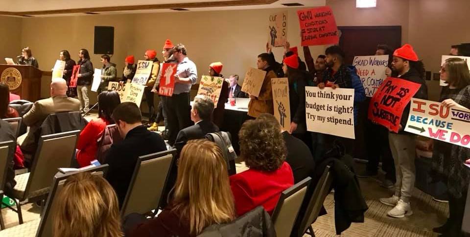 In the background, about 15 ISU students holding up various signs seem to be interrupting a formal meeting, some wearing the red hats of the graduate students union. Most of the students are looking at a person speaking at a podium. One sign reads: ‘You think your budget is tight? Try my stipend.’ Another reads ‘ISU WORKS BECAUSE WE DO.’ Several signs have imagery of the power fist holding a pencil. In the forefront, we see the backs of individuals seated and facing the podium.