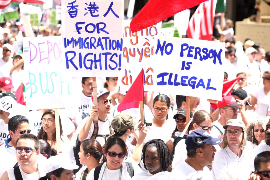 A diverse crowd of people are packed togetheron a sunny day. They hold signs reading "for immigration rights" and "no person is illegal."