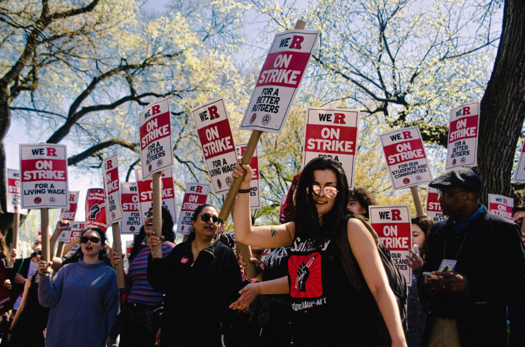 A celebratory crowd with a feminine person in sunglasses dancing in front holds signs reading We R on Strike for a Better Rutgers.