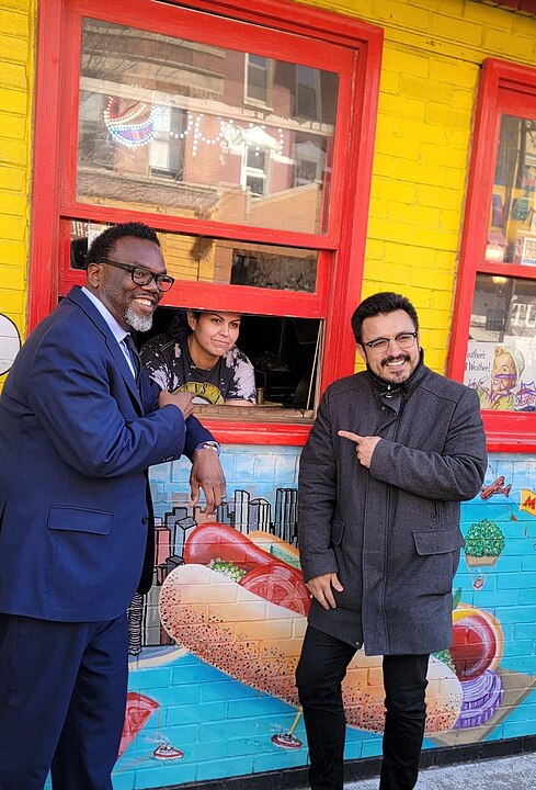 Democratic Socialist of America member, and Chicago Alderperson, Byron Sigcho-Lopez smiling and pointing enthusiastically at Chicago Mayor-elect Brandon Johnson, while standing in front of a hot-dog stand, with vendor in the picture. Taken in March 2023.