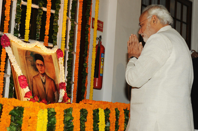 Indian Prime Minister Narendra Modi, with hands held in prayer and head bowed,over a photo of Veer Savarkar, a Hindu nationalist leader and supporter of European fascism.