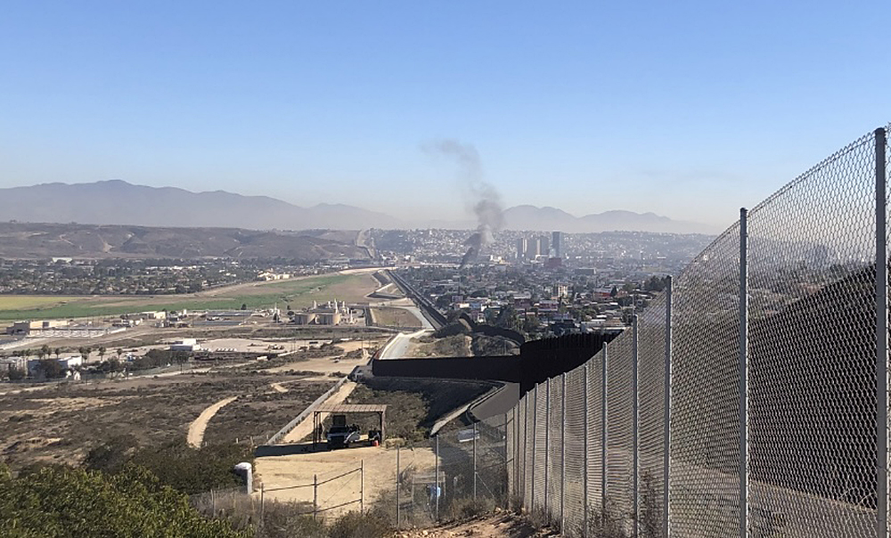 Landscape shot looking down the border fence that goes up and down rolling hills. On the left of the fence is open space and a residential area, and on the right is a dense urban area with a number of tall buildings.
