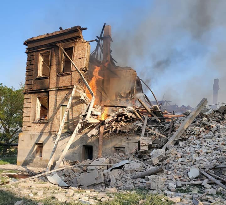Orange flames rise out of the ruins of a brick and concrete school. Only one corner of the building is still standing, and there’s a pile of rubble to the right of it.
