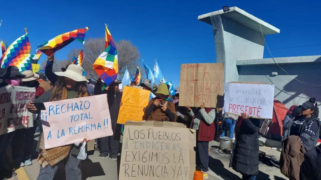 More than a dozen of protesters in hats stand in the sun waving multi colored banners and carrying signs reading Rechazo Total a la Reforma! Los Pueblos & Indigenas Exigimos la Renuncia," and other slogans.