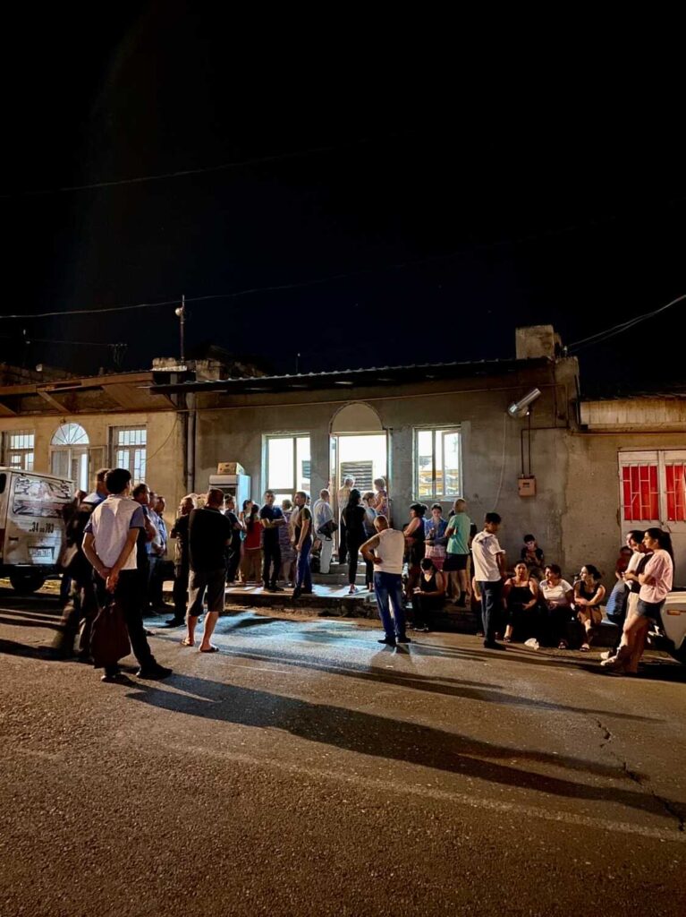 A group of people mills about in front of a lighted storefront in the hours before dawn. They are Armenians waiting in a breadline during a blockade.