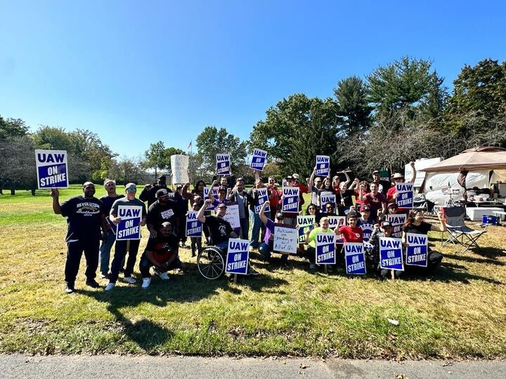a group of about 40 people holding UAW ON STRIKE picket signs, sitting and standing on a sunny hillside.