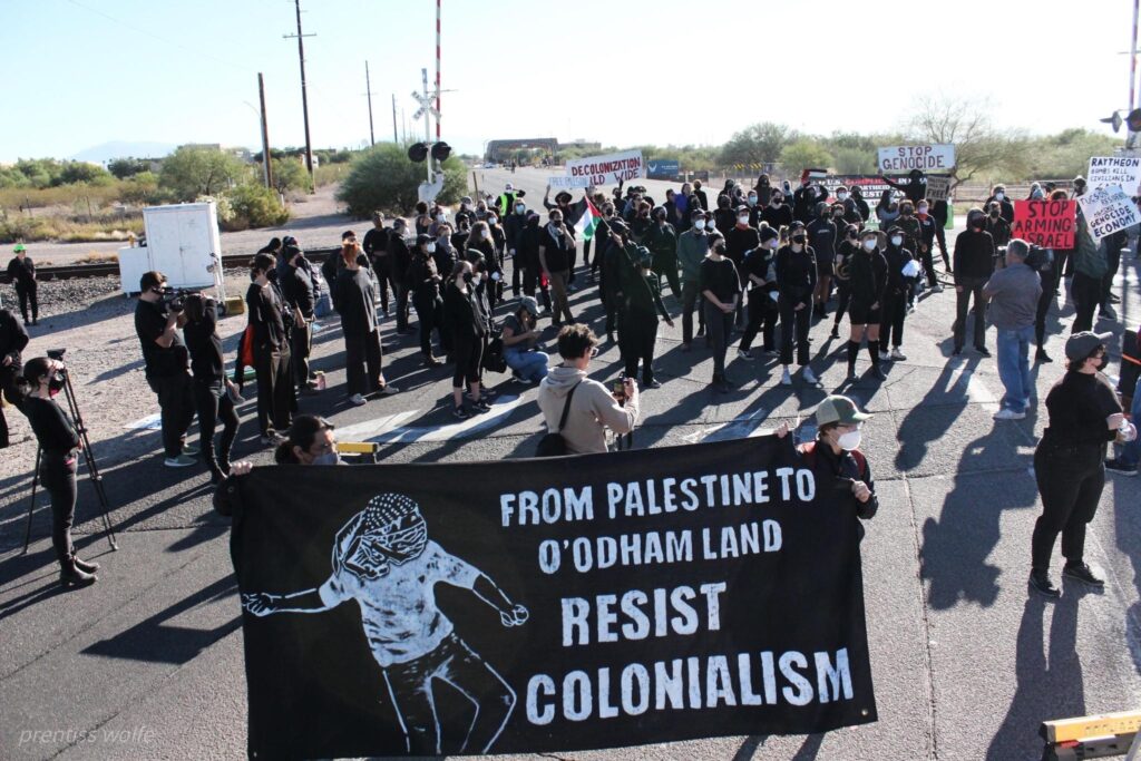 About 60 masked activists wearing black are gathered on pavement near railroad tracks. In the foreground, two hold a banner, reading, in white on black, "From Palestine to O'Odham land: Resist Colonialism." There is an image of a figure throwing a rock.
