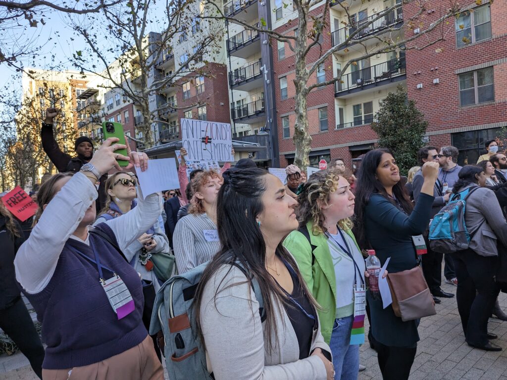 Dozens of protesters gathered to listen to speakers in solidarity with Palestine with fists raised in air. One sign reads Anti-Zionism does not equal Anti-Semitism.