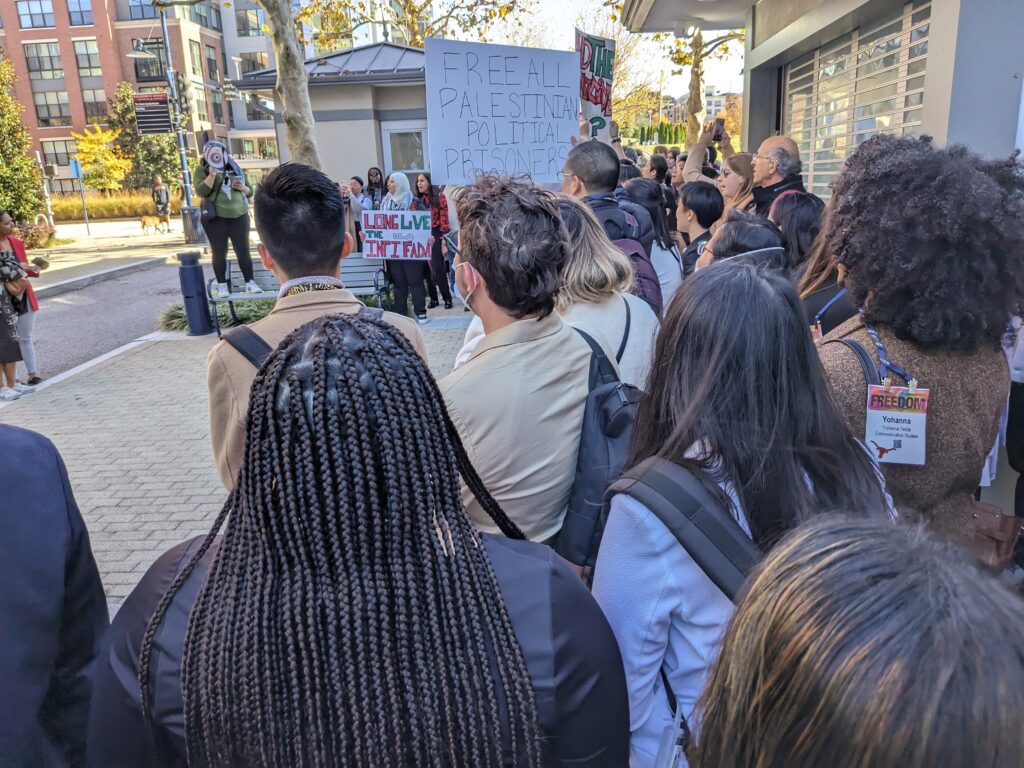 Protesters, photographed from behind, listen to a speaker standing on a bench speaking into a megaphone. Signs read Long Live the Intifada and Free all Palestinian Political prisoners.
