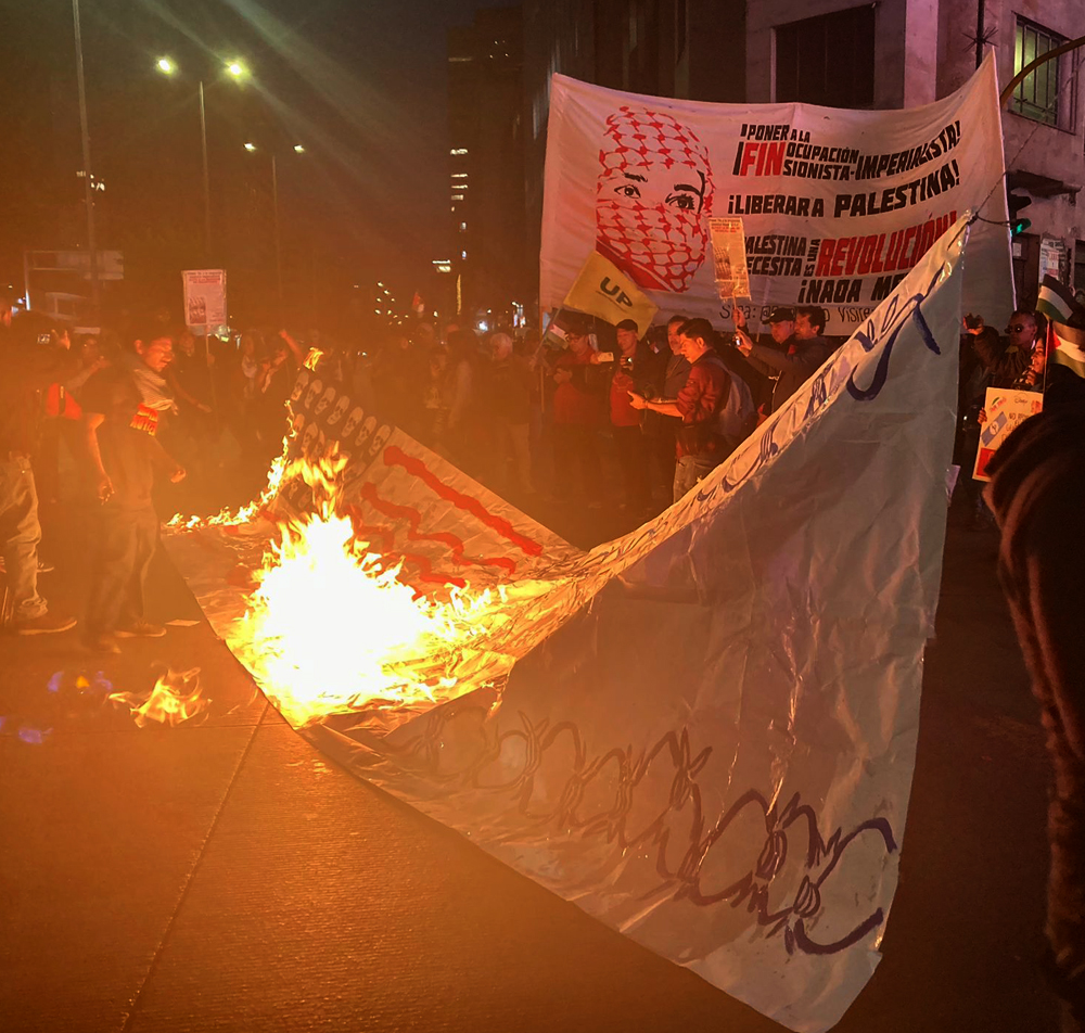 Nighttime crowd shot on the street. The middle of the photo shows a large a hand-painted banner in flames. The banner has red stripes—like an American flag—with skulls where the stars would be, plus barbed wire. In the background is a banner in Spanish with a head figure in a red keffiyeh and the slogan ¡Liberar a Palestina!
