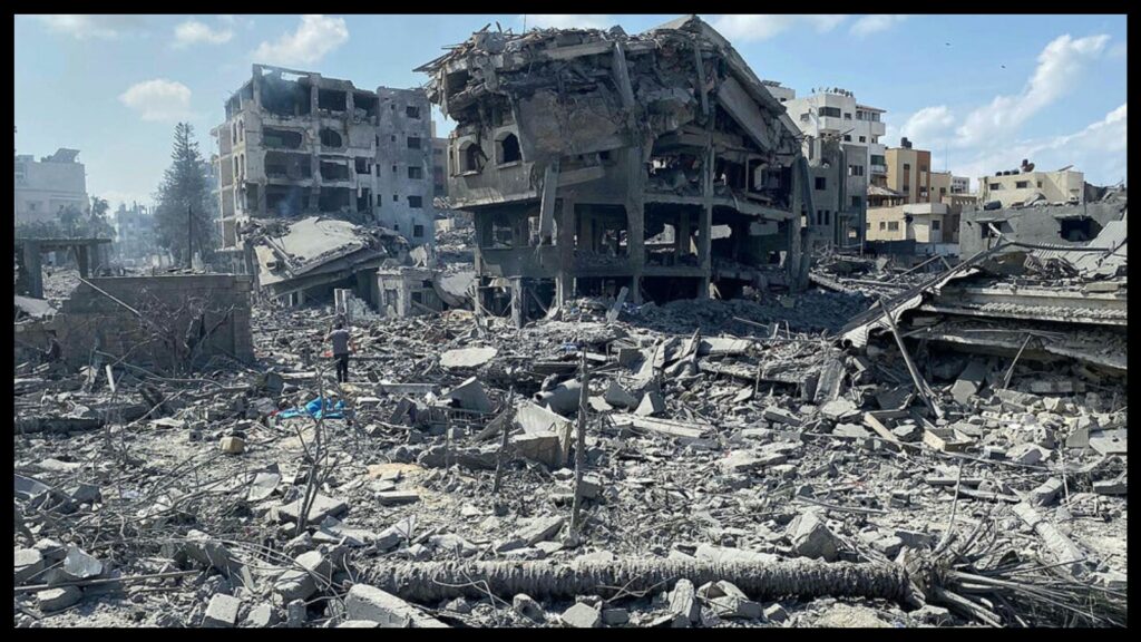 In the foreground is gray urban rubble from bombing. In the middle distance are damaged buildings. The upper two stories of the building in the center has collapsed on the lower two stories, and the structure looks like it could collapse entirely at any moment. There appears to be some dust in the air, but the sky behind is blue with scattered clouds