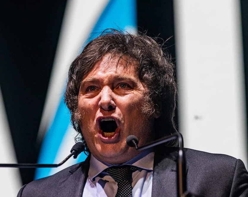Photo of newly-elected Argentinian President Javier Milei with his trademark long hair and side-burns, mouth wide open as though screaming into two microphones framing his face.