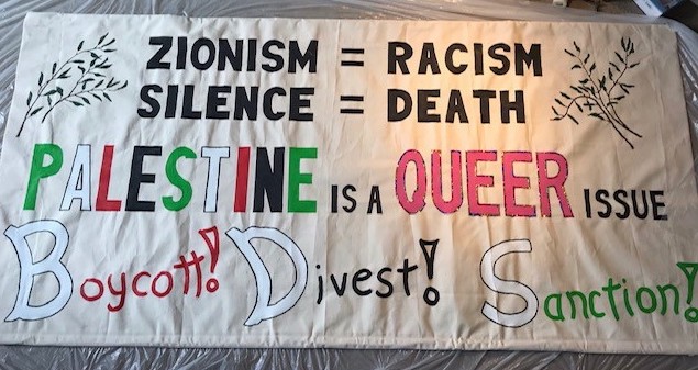 Banner of NYC Queers Against Israeli Apartheid, circa 2019, which reads, in black lettering at the top, “Zionism equals racism” and just below, also in the same black lettering, “Silence equals death.” Across the middle of the banner it reads: “Palestine is a queer issue.” The word “Palestine” is written in alternating white, green, red, and black of the Palestinian flag. The word “queer” is written in pink lettering. The bottom of the banner reads: “Boycott,” “Divest,” “Sanction” with each initial letter in white, with the rest of the first word in red, the rest of the second word in black, and the rest of the third word in green. Images of olive branches decorate the upper left and right corners of the banner.