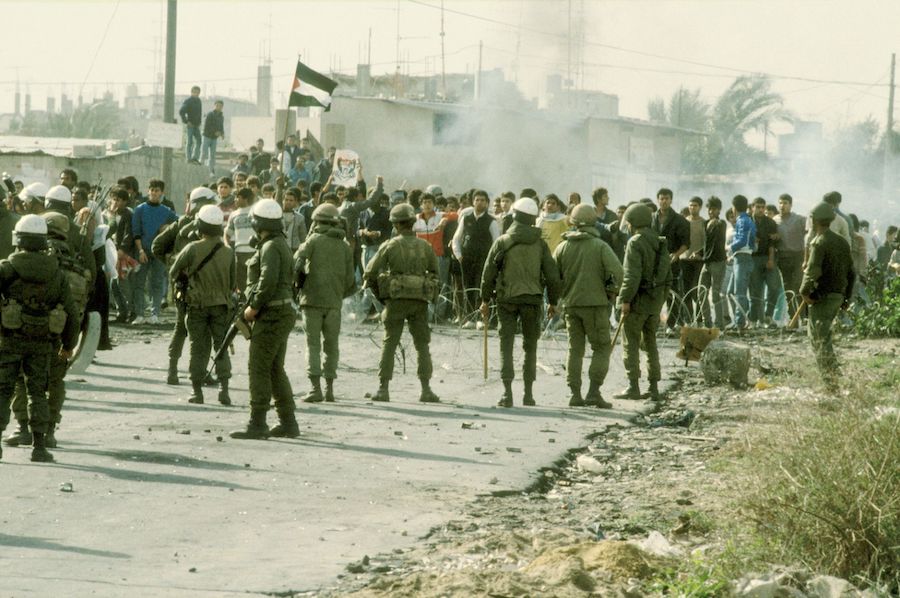 Color photo on a rubble-strewn road with weeds off to the right. The shot is taken from behind about a dozen soldiers, many with white helmets. They carry truncheons and assault rifles. Facing them across a razor-wire barrier is a crowd of fifty or more unarmed young people, apparently all boys. Smoke rises just behind the crowd. One protester holds up a picture that appears to be of Yasser Arafat; another holds up a Palestinian flag. Some distance back, two boys stand on a the wall of a crude one-story structure. One of them has a posture as if he is following through on throwing a rock toward the soldiers.