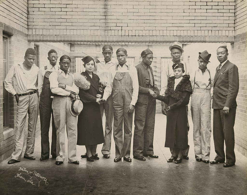 A black and white photo of 11 Black people in a hallway in the 1930s. They are the 9 Scottsboro Boys, Juanita Jackson Mitchell, and Laura Kellum. They are wearing or carrying tats and dressed variously in casual pants, overalls, and suits.