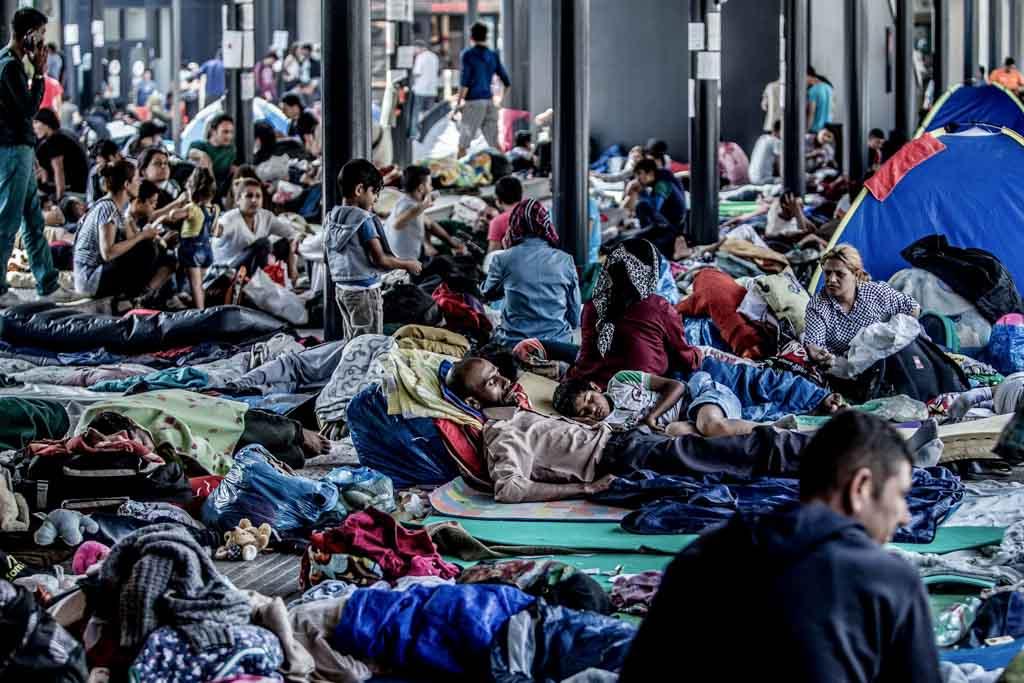 Many people in a dark room lay on the floor, slouched against walls, sleeping men, women, and children. Some children are walking around.
