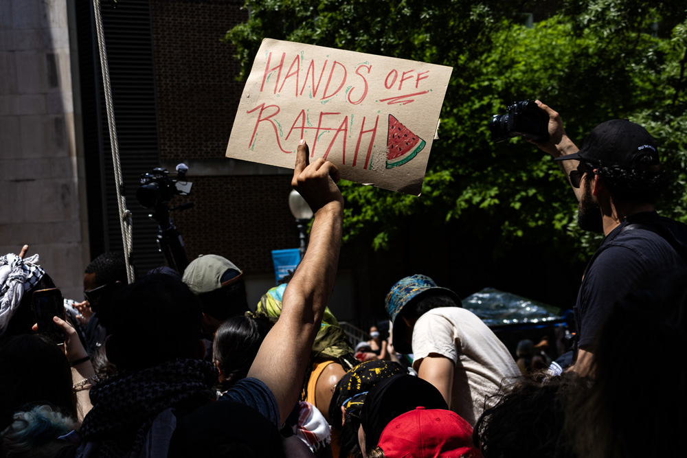 Dark image of a crowd with one hand raised above holding a hand-lettered sign saying “Hands off Rafah.” Sign also contains a depiction of a green and red watermelon slice.