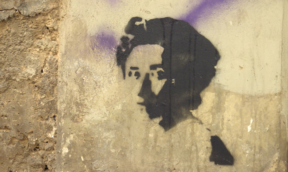 Stenciled image of the head and shoulder of a woman in three-quarter profile. The image is in black spray paint on a yellow concrete wall.