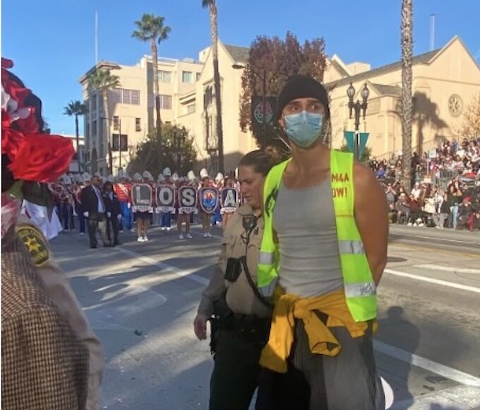 Palestine solidarity march participant, under arrest at the Rose Parade. Participant wearing a fluorescent yellow safety vest and mask with hands handcuffed behind his back being led from behind by a police office in tan and brown. In the background are the march participants carrying the Palestine banner (not legible). Photo by Teto.