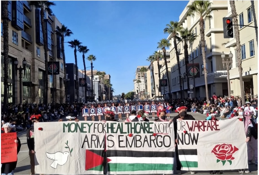 Palestine solidarity protesters carrying a banner at the Rose Parade on January 1, 2025. White banner with a painted Palestinian flag at bottom (red, green, white, and black), a painted white dove in left corner and red rose in right corner. Banner reads, in green, red, and black lettering, “Money for Healthcare not for Warfare; Arms Embargo Now.” Photo by Teto.
