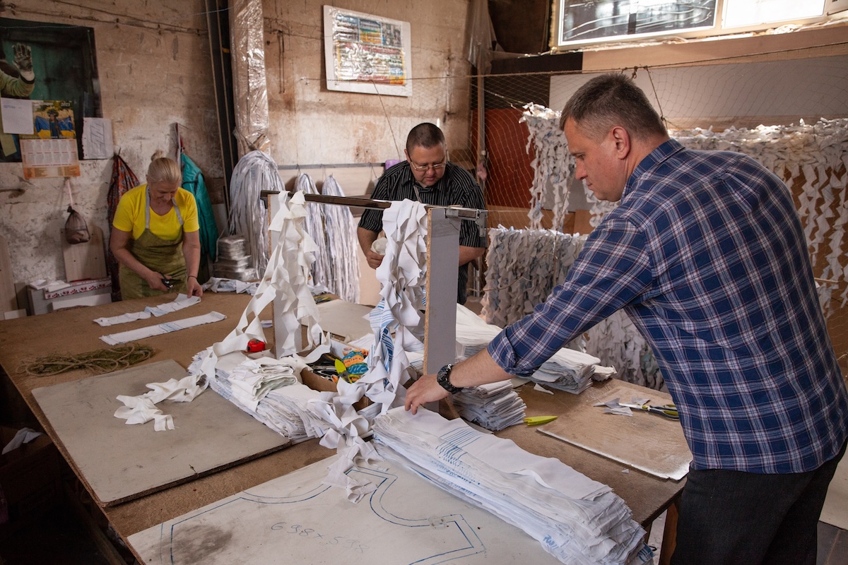 Volunteers in Zaporizhzhia make camouflage netting, photo shows three adult volunteers working around a table with scissors and fabrics. This location was struck by Russian missiles earlier this year. Summer 2023. Photo by Kris Parker