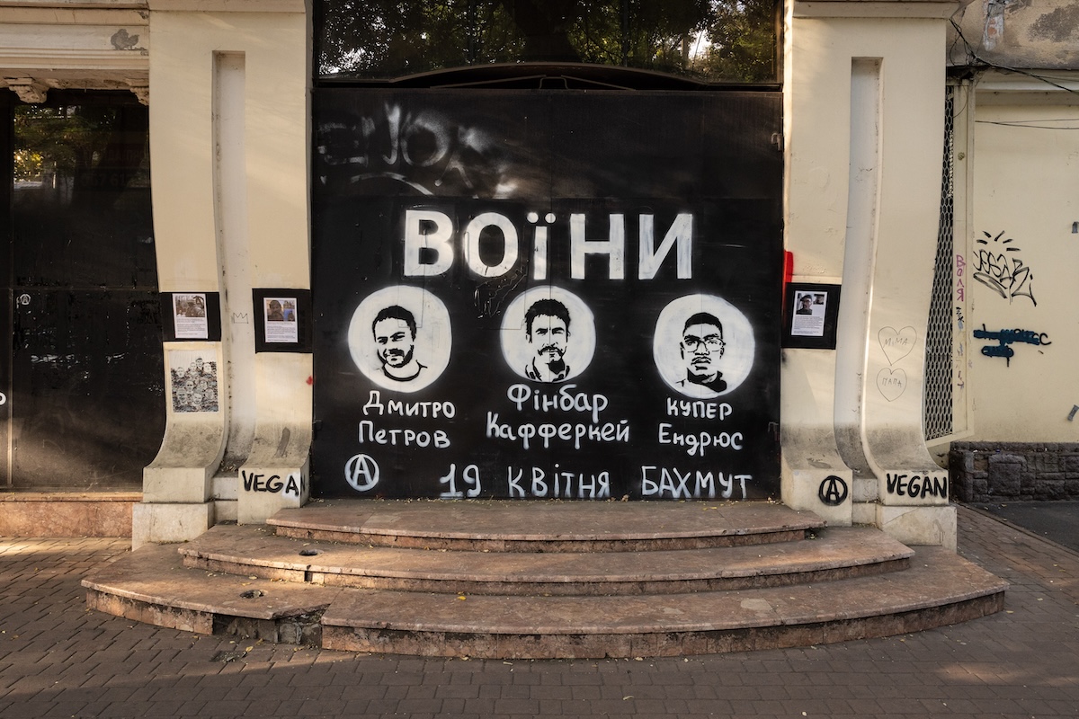 A memorial in Odessa for three leftist soldiers killed defending Bakhmut in 2023. A black banner with white silhouettes of Dmitry Petrov of Russia, Finbar Cafferkey of Ireland, and Cooper Andrews of the U.S. during the summer of 2024. An “A” anarchist symbol in the bottom left corner. Photo by Kris Parker.