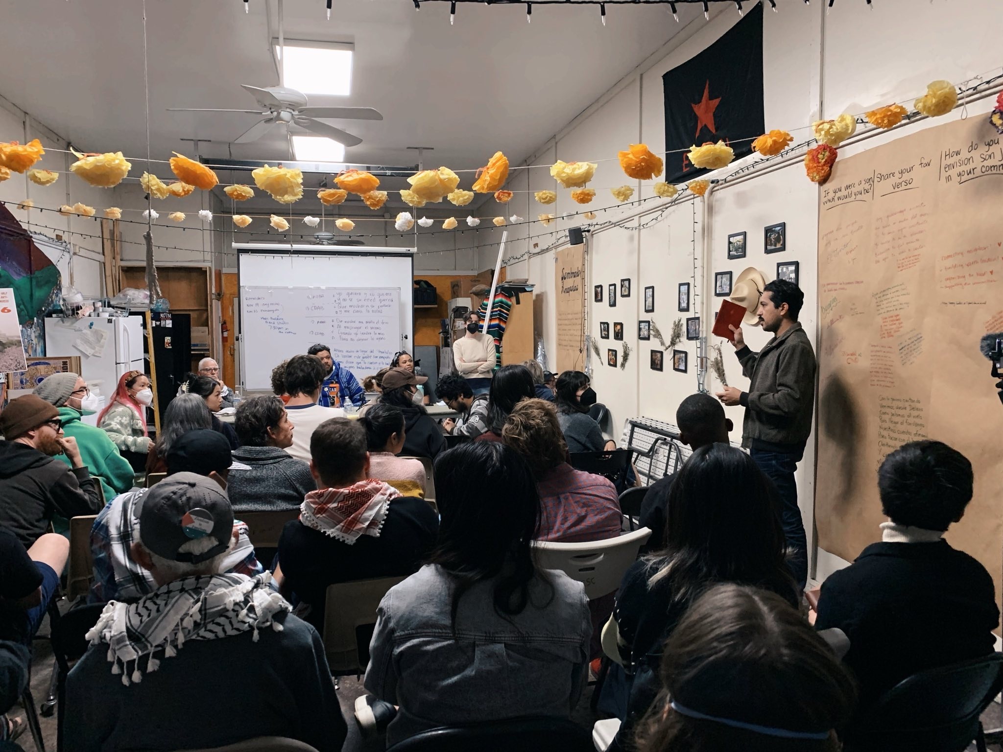 A crowd of people sits in front of a panel with a presentation. A person stands to the right of the room speaking and holding a book.