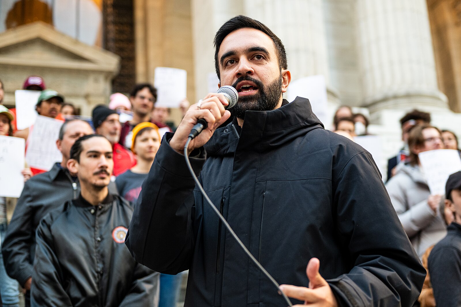 Foreground: Bearded young man speaks speaks outdoors into a microphone. Background: Rally-goers hold signs and listen as they stand on the steps of a building with large columns.