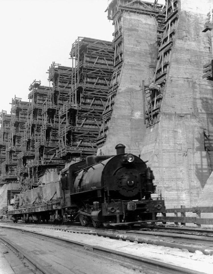 Massive concrete columns being built with wooden scaffolding, with a steam locomotive in the foreground.