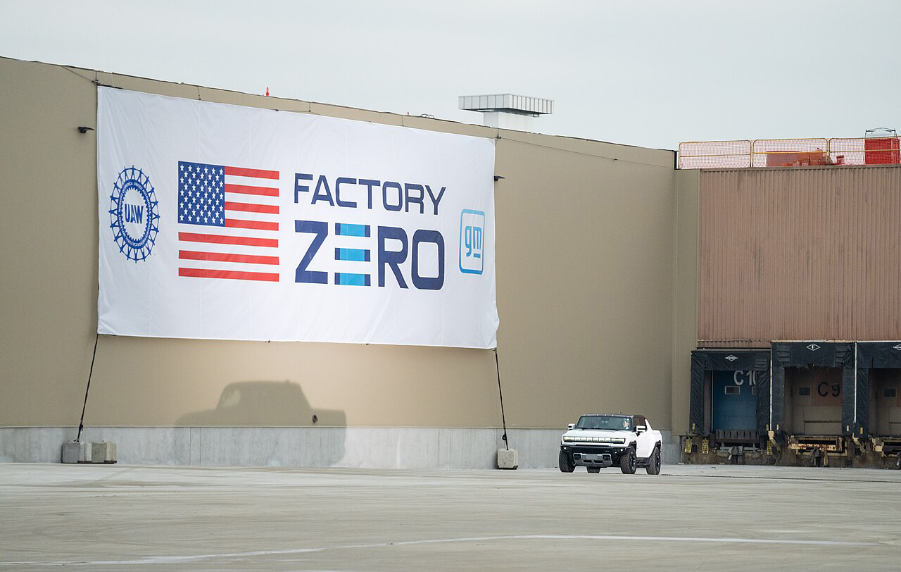 Huge banner outdoors on a blank wall has a U.S. flag, the logos of GM and the UAW, and the words, “Factory Zero.” In front of that is a white Hummer.