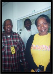 A photo taken at a correctional facility, showing, from left to right, Walter Reed, now deceased, the father of Rodney Reed dressed in a black and red checked flannel shirt, Rodney Reed (center), in a white overcoat, and Sandra Reed, in a yellow t-shirt with red lettering and a black sweater.