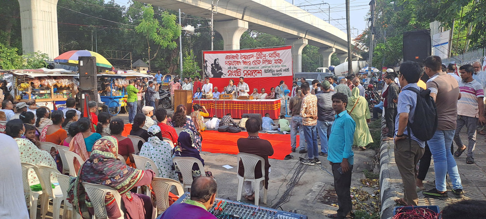 Wide shot of a street rally, from behind a crowd mostly seated on plastic chairs. Toward the front, children are bent over artwork they are creating, and beyond that is a speakers’ table, draped in red and gold, with a wide banner behind. Most of the banner is in Bengali, with “Stop genocide. Ceasefire in Gaza now,” in English.