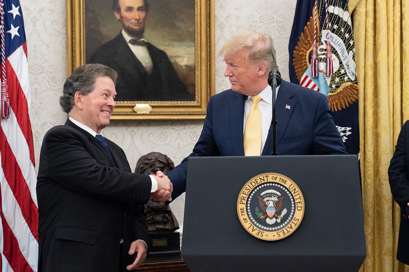 President Donald Trump stands at a podium in front of a portrait of Abraham Lincoln, smiling and shaking the hand of economist Arther B. Laffer.