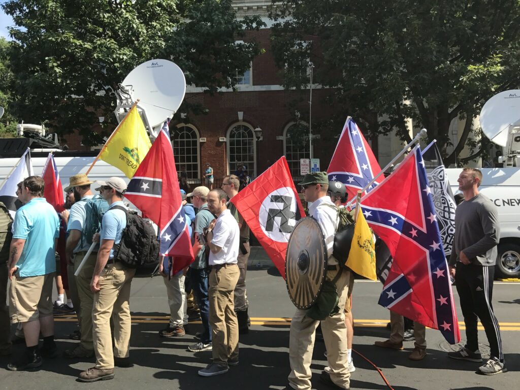 About a dozen white men wearing khaki pants carry Confederate, Nazi, and Gadsden (don't tread on me) flags in a sunny street. News vans in background.