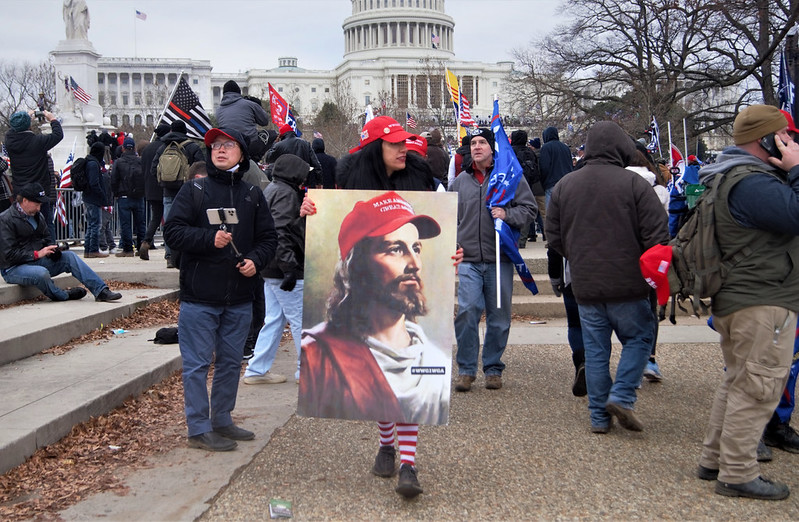 In front of the U.S. capitol in winter wear, about two dozen protesters in hats. One woman in foreground holds a painting of Jesus Christ wearing a red "Make America Great Again" hat.