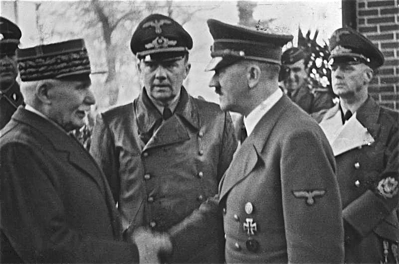 Black and white photo. on the left wearing a military hat and suit is an older man who is Philippe Petain; On the right shaking his hand is Adolf Hitler in military uniform. They are surrounded by other Nazis.