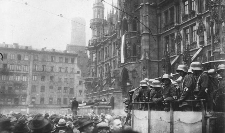 Black and white image of crowd on a city street with Nazi troops on balcony above. In the background, a man is elevated and speaking to the crowd.