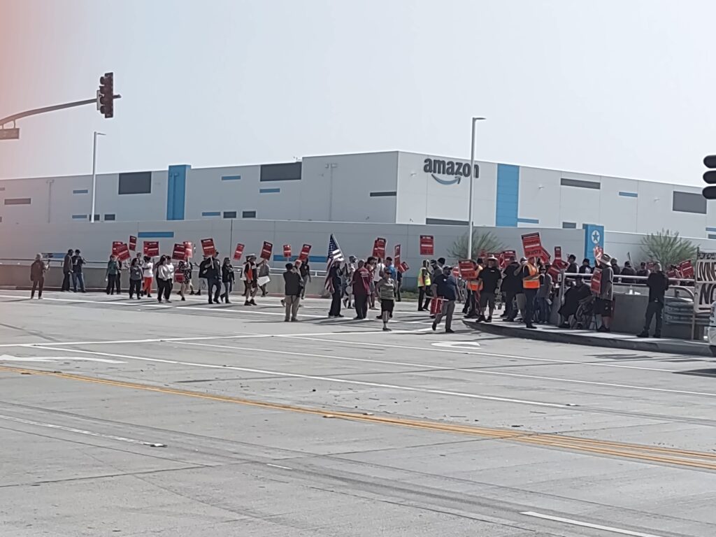 About three dozen people march in front of a large Amazon warehouse carry red picket signs reading "Amazon Prime Beware/Amazon Air is Unfair," and one picketer carries an American flag.